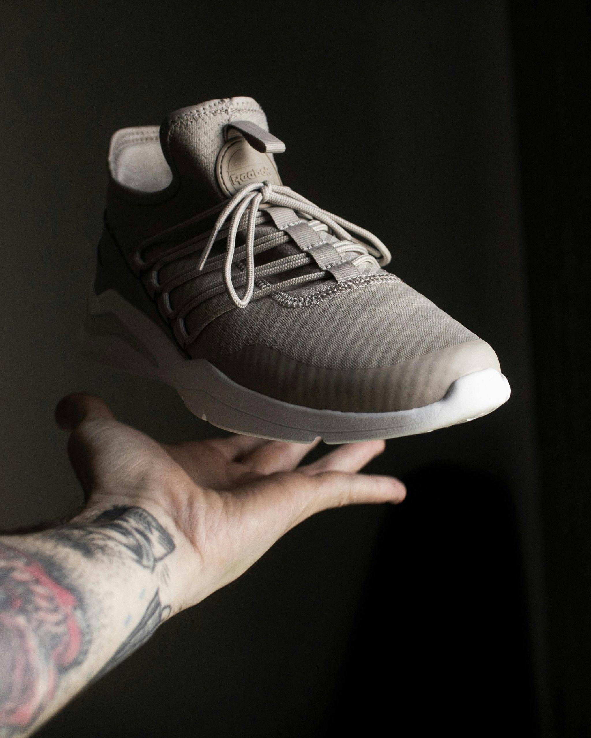 Close-up of a beige sneaker floating above a tattooed hand in low light.